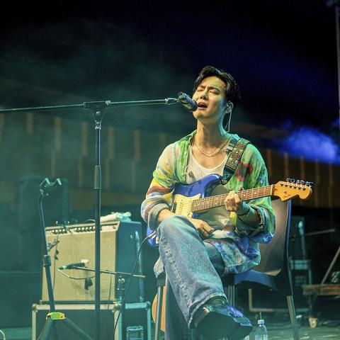 SUHO sits on a chair, singing into a microphone, with his blue and white guitar on his lap. He is wearing blue jeans and has one leg crossed over the other. His shoes are chunky. He has a white tank top on under an unbuttoned long sleeve loose fit shirt in greens, blues, reds water colors.