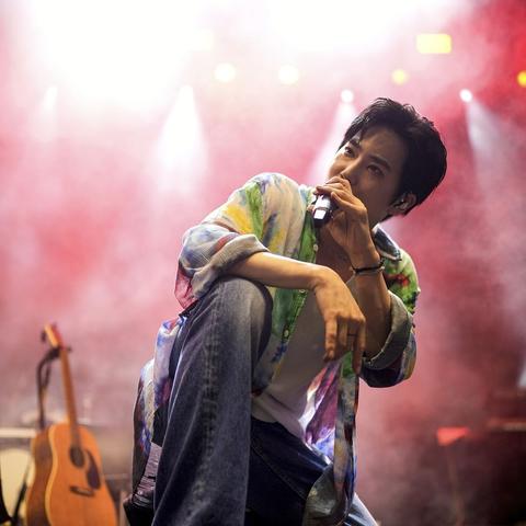 SUHO crouches on stage, singing into a microphone with stage lights glowing through smoke behind him. He's wearing blue jeans and a white tank top underneath a loose unbuttoned water color wash dyed shirt with greens, yellows, blues, reds, and purples.