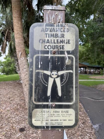 A post with a sign that reads: “Advanced Timber Challenge Course”
The panel below is black with an image of a person bent at the waist with their arms to the side at 90°.
The bottom panel describes how to do a lateral arm raise, and suggests you should perform this activity either 10 or 20 times at this spot, depending on your fitness level. 
In the image of the person, where the head is, someone has stuck on 2 googly eyes.

The sign is on a brown post next to a paved walkway, surrounded by areas of grass, mulch, and trees. There is a picnic canopy blurred in the background.