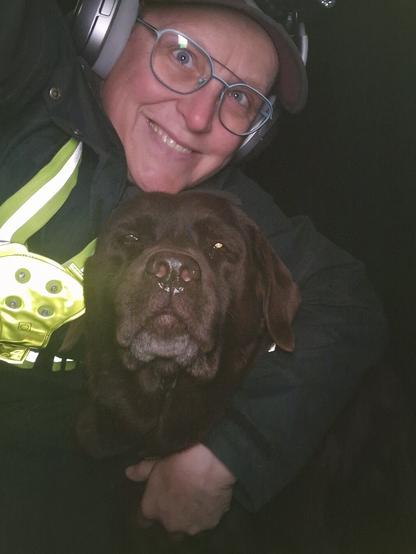 Night time selfie of a person and a chocolate brown Labrador with greying snout. The person is wearing a black jacket, yellow safety harness, red cap, silver headphones and blue-rimmed glasses. On their cap rests a headstrap torch in off position. Their free hand is around the dog's shoulders. Both are facing the camera. The person's head is resting on the dog's head. They're smiling, grateful for their time spent together.