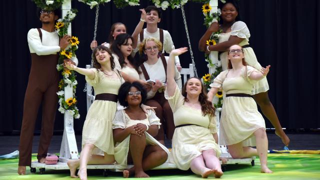 A group of young performers is posing on stage, dressed in light-colored dresses and brown outfits. They are arranged around a white swing adorned with flowers, featuring sunflowers and daisies. The scene conveys a cheerful and lively atmosphere.