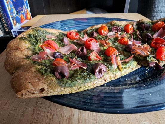 A close-up cross-section image of several slices of homemade pizza. Whole-wheat dough has given the pie a more rustic color and texture than is typical with commercial pizza. Distinct air bubbles can be seen through the cross-section of dough. It is topped with pesto, olives, cherry tomatoes, and prosciutto. The image is heavily filtered in the style of Instagram.