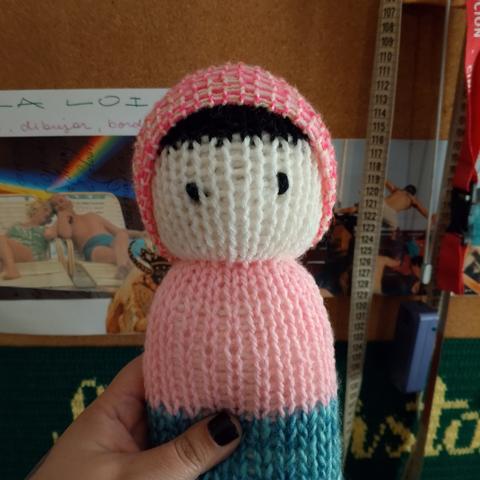 Mano sosteniendo a un muñeco tejido en punto, tiene un gorrito rosa, pelo negro, ojos negros, cara blanca, ropa rosada y azul y zapatos rosados.