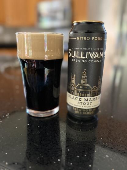 A pint glass full of dark beer with a thick tan head. The black can has a cityscape in gold and lettering in white and gold