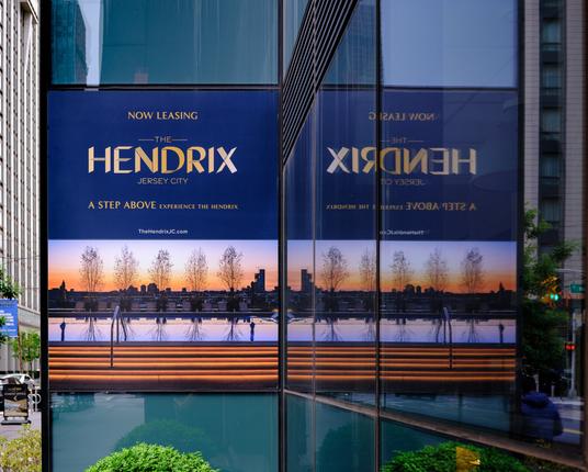 A sign advertising available leases in a building called "The Hendrix": NOW LEASING / THE / HENDRIX / JERSEY CITY / A STEP ABOVE experience the hendrix.  There are various amusing reflections and colors in the photo.