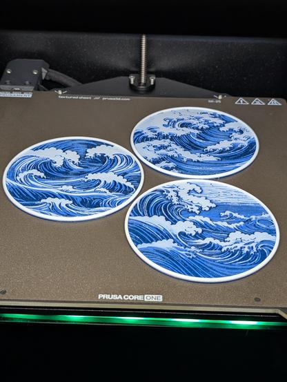 A 3D printer bed with a green light under it. There are 3 coasters on the print bed with blue waves on them with white caps and clouds