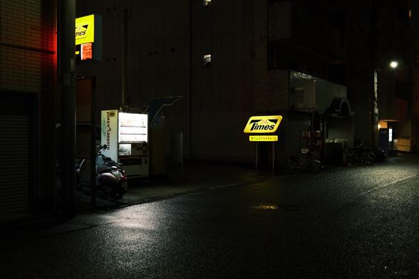 A yellow lit sign advertises 24 hour parking on a rainy night. The pavement shines with the light rain reflecting the light from signs and nearby streetlights.