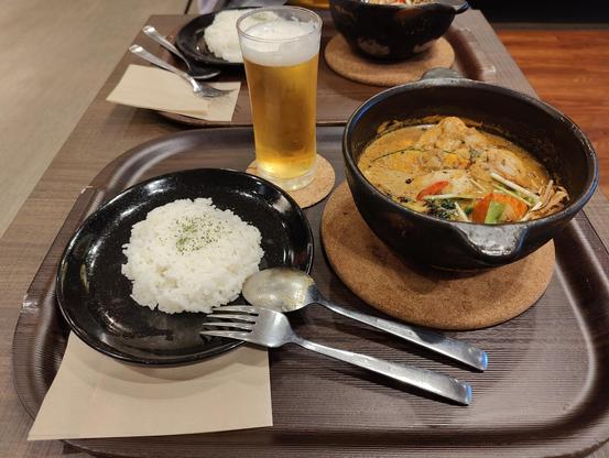 A photo of a meal set. You see a big bowl of orange curry filled with meat and vegetables, plus a side of rice and a small beer.