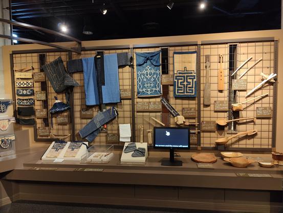 A photo of a museum exhibit. On the wall you see various pieces of Ainu clothing and wooden cooking tools.
