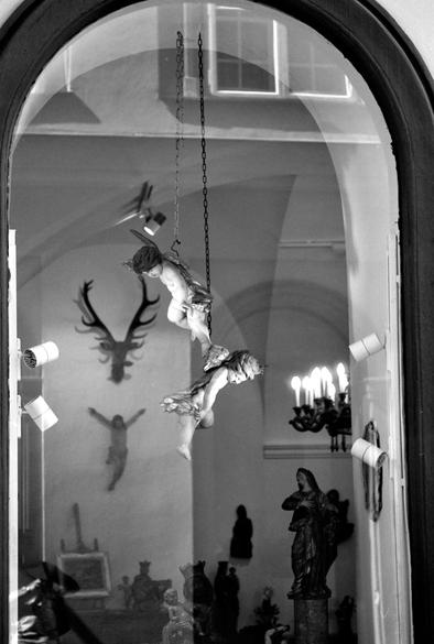 The black and white image shows two cherub-like angel sculptures suspended by chains inside a room filled with religious statues and artifacts, with reflections and shadows adding a surreal atmosphere.
