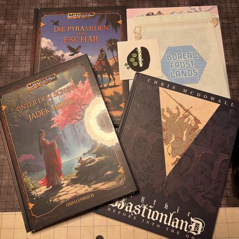 Die Folgenden Bücher liegen auf einem Tisch: “Midgard 5 - Unter dem Schild des Jadekaisers”, “Midgard 5 - Die Pyramiden von Eschar”, “Boreal Frostlands” und “Chris McDowell - Mythic Bastionland”