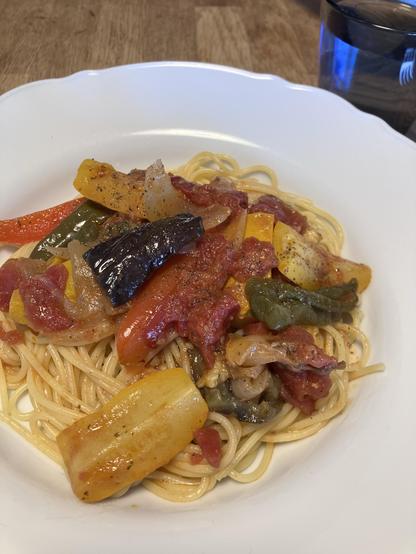 A plate of spaghetti topped with a colorful mixture of sautéed vegetables, including bell peppers, onions, and tomatoes. The dish is seasoned and served in a white bowl, with a glass of water in the background.