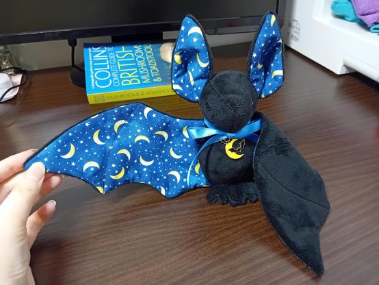 A black plushie bat with big ears. A dark blue cotton lining covered in yellow crescent moons is used for the front of the wings and ears. It also has a blue satin ribbon and a moon charm around its neck.