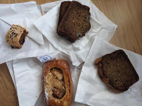 4 torn white paper bags with food on top of it.  The food is a mini sausage roll, 2 X slices of banana loaf, a single slice of banana bread and an apple pastry.