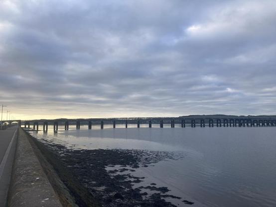 View across Tay Firth along the flood wall toward the rail bridge a strip of silver morning light breaks the various shades of steely blue