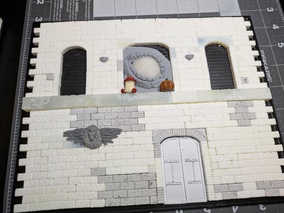 A model brick wall with arches on two floors. The ground floor now has an unpainted plastic door with details glued and scratched on. The middle arch on the second floor has a giant reptilian eye looking in, with a pumpkin monster and mushroom monster sitting in front of it