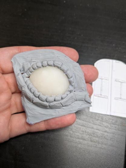 A gray polymer clay dragon eye with sculpted scales around it. It's in the shape of the archway and held in a hand