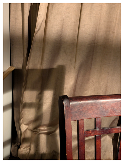 A dark wooden chair with a slatted backrest sits in front of beige curtains and cast a shadow.