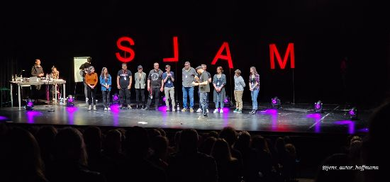 Das Bild zeigt die Bühne des Staatstheaters Augsburg im Martinipark beim Finale der Bayerischen Slam-Meisterschaften 2023. Im Hintergrund leuchten groß die Buchstaben „S L A M“, von hinten beleuchtet in kräftigem Rot, das "L" spiegelverkehrt.

Auf der Bühne stehen rund ein Dutzend Menschen – die Finalist*innen des Abends, Slammer*innen aus ganz Bayern, die sich zuvor über Vorrunden für diesen Höhepunkt qualifiziert haben. Sie stehen nebeneinander in einer Reihe, einige lächeln ins Publikum, andere schauen konzentriert. Links im Bild ist ein DJ-Pult mit zwei Personen zu sehen, von wo aus der Abend musikalisch begleitet wird.

Ganz vorne, gut sichtbar, steht Moderator Horst Thieme, erkennbar an seiner Schiebermütze. Er spricht gerade ins Mikrofon, während das Publikum – angedeutet als dunkle Silhouetten im Vordergrund – gespannt zuhört. Die Atmosphäre wirkt feierlich, lebendig und gespannt – gleich wird der Sieg verkündet.