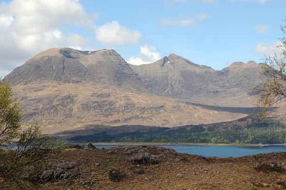 Beinn Alligin in Wester Ross. The image shows a view over a loch to high mountains that rise to three connected peaks. There are small patches of snow on near two of the summits. In the foreground is the rocky near side of the loch. The mountains are in sunlight.