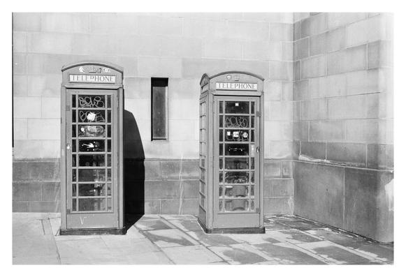 his black-and-white photo shows two traditional British telephone booths side by side against a stone wall. Each booth has a rectangular shape with a domed top, featuring the word "TELEPHONE" and a crown symbol above the door. The glass panels reveal some graffiti and stickers inside, with phrases like "LOVE" and "SOHO" visible. Shadows from the booths stretch across the pavement, suggesting sunlight. The wall behind is made of large, rectangular stone blocks, with a narrow, vertical window between the booths. The scene captures an urban setting with a hint of wear and character.