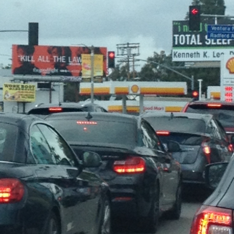 Lines of all black cars with red illuminated brake lights stopped at a traffic light with a sign that says "Ventura" above "Radford". The background is cluttered with poles, trees in front of a cloudy gray sky, a Shell station, and signs. The only words visible on the signs are "work boot", "kill all the law" and "total sleep", adding to the bleak dystopian feeling of the picture of mundane LA traffic