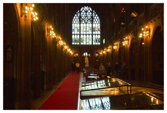 Main corridor of the John Rylands Library in Manchester. In the middle there are some tables and chairs. On the left a red carpet goes from back to front and at the end of the carpet there are two people walking away. On the Background a giant decorated window with tracery and a white statue right under it. On the left and right sides there are some arcs leading to smaller quieter areas.  On the right side there's a group of people waling towards the camera
