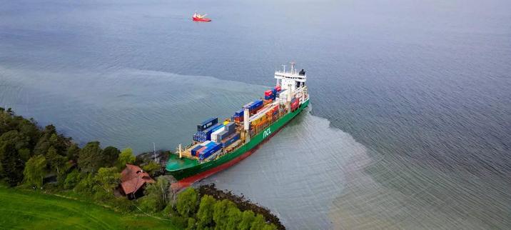 Big ass cargo ship with loads of containers stuck nose first a few meters away from a hut at a Norwegian fjord.
