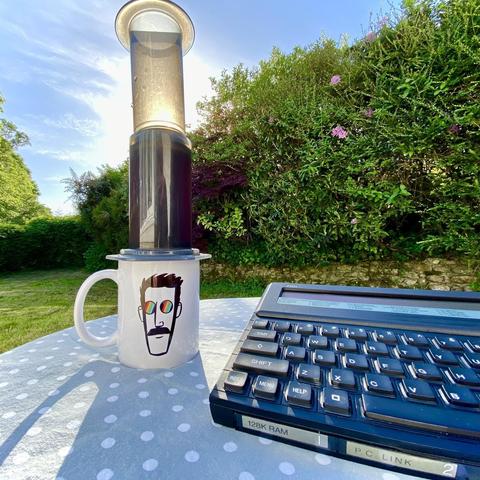 Photograph, outdoors, garden. In the foreground a WhatHoSnorkers coffee mug with an Aeropress atop, such that it looks like Snorkers is wearing a top-hat. (Well sort of). The mug is on a little table and is next to a Cambridge Computer Z88 portable computer. The sun is rising in the background. It is evidently Snorking Time™️.