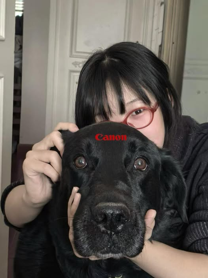 Una persona con rasgos asiáticos está ligeramente recargada sobre un perro grande color negro, sosteniendo su cabeza como si fuera una cámara fotográfica: su mano izquierda sosteniendo el hocico por debajo, como si fuera la lente, su mano derecha sosteniendo la oreja y en posición de pulsar el botón de disparo, en su frente, aparece sobrepuesto el logotipo de la marca Canon. El perro mira tranquilamente a la cámara que toma la foto.
