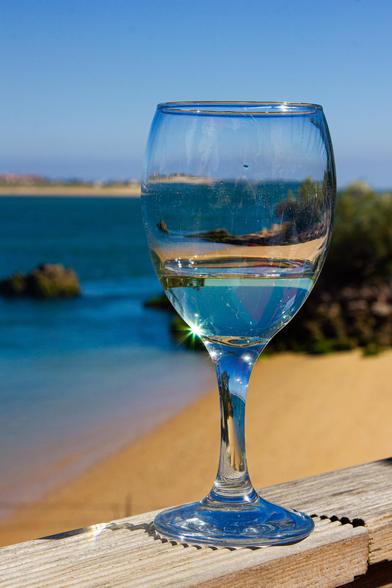 In the middle of the photo a glass of wine on a wooden beam (from the conversion of a terrace) with a little white wine in it. The sun gives a star on the edge of the glass. In the background, vaguely, a beach with sea and blue sky.