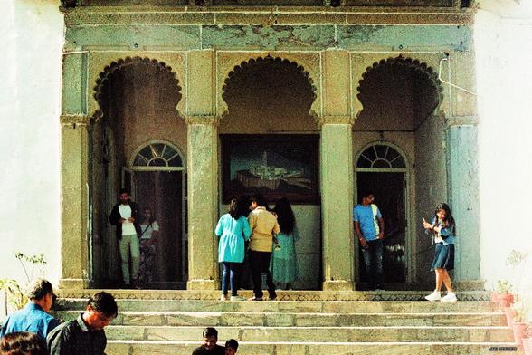 Foto in Retrofarben, aufgenommen an einem Eingang zum Hauptgebäude des Monsunpalastes bei Udaipur, Rajasthan, Indien. Eine Treppe führt durch drei auf Säulen gestützte Bögen ins Innere. Im Vordergrund, auf der Treppe und im Eingangsbereich halten sich Besucher in indischer und westlicher Kleidung auf. Leica R4 mit 50 mm Summilux auf Rollei Crossbird-Film.