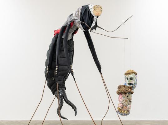 Image: sculpture of what looks like a very large black earwig with thin steel-bar-like legs and pretty terrifying pincers at the bum end; at the head end, we seem to see a pinned-on photo of a pale human face with too many eyes; the upper body seems to be wearing some sort of messed-up suit; a strange object appears to be attached to one of the critter’s legs – one or more heads made of strange material, papier-mâché or sweets or coloured cotton balls or…; this all seems to be in some sort of gallery space • “Curated by Sara Greavu ( Project Arts Centre) in 2023, Kian Benson Bailes’ Culchie boy, I love you / Grá mo chroí thú, mo chábóigín féin arrives at Mermaid Arts Centre in a new iteration. Comprising sculpture, digital & material collage, musical instruments, ceramics, textiles, woodwork, and live sculptural sound elements. Culchie boy, I love you / Grá mo chroí thú, mo chábóigín féin references Irish folklore, mythology, and craft traditions in an exuberant expression of rural…”
