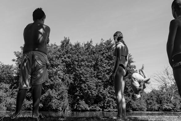On the banks of a river, three teenagers watch as a third does a somersault to dive into the water. His head is down and his body is bent, blurred.