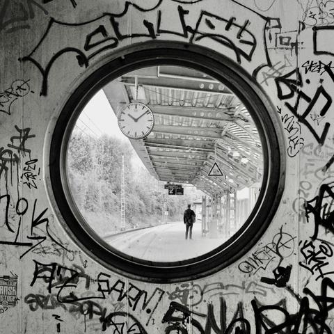 Das Bild zeigt einen Bahnsteig durch ein rundes Fenster in einer mit Graffiti bekritzeln Wand. Die Graffiti sind abstrakt, die Tags kaum zu entziffern. Durch das Fenster sieht man einen Bahnsteig mit einem modernen, metallenen Dach. Eine analoge Bahnhofsuhr ist sichtbar und zeigt 10:09. Im Hintergrund sind Bäume neben dem Bahnsteig zu sehen. Auf dem Bahnsteig, der sich in die Ferne erstreckt, steht eine einzelne Person mit dem Rücken zum Betrachter. Er scheint, den Bahnsteig entlang zu schauen – möglicherweise auf eine Anzeige. Die Person trägt dunkle Kleidung und einen Rucksack. Die Atmosphäre ist still, einsam und etwas melancholisch, mit einem Kontrast zwischen der rauen, urbanen Ästhetik der Graffiti und der ruhigen, fast stillen Szene auf dem Bahnsteig. Das Foto vermittelt ein Gefühl von Isolierung und dem Warten auf etwas.

The picture shows a train platform through a round window in a wall scrawled with graffiti. The graffiti is abstract, the tags barely decipherable. Through the window you can see a platform with a modern, metal roof. An analog station clock is visible and reads 10:09 and in the background you can see trees next to the platform. On the platform, which stretches into the distance, a single person stands with their back to the viewer. The person appears to be looking along the platform - possibly at an information display. The person is wearing dark clothing and carrying a rucksack. The atmosphere is quiet, lonely and somewhat melancholic, with a contra