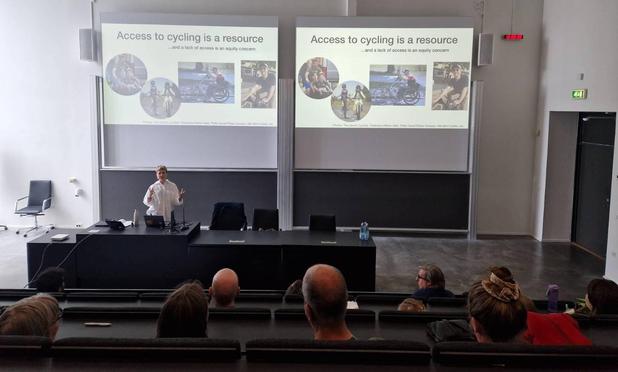 Photograph of a lecture hall, with female researcher giving a presentation at the pult. Three rows of heads from the audience visible from behind. The slide is titled "Access to cycling is a resource" showing different examples of cyclists.