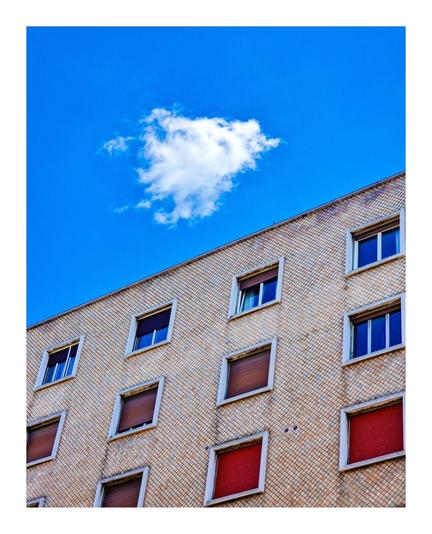 The photo is of the top of a building with three rows of square windows with a vibrant blue sky above it. It is made of light-brown bricks and is slanted - dipping towards the left of the photo. The windows are in a grid pattern and like the building they are slanted. There are multiple windows, each with a white frame and red or blue shades. The windows are rectangular in shape and have white frames. The sky above the building is a bright blue, with a single white fluffy cloud.