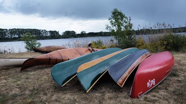 Foto von sieben Kanus die meist kopfüber, nebeneinander an Land liegen. Im Hintergrund Gestrüpp, ein See und bewölkter Himmel.