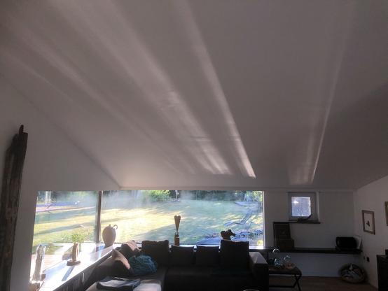 Das Bild zeigt einen modernen Wohnraum mit einer schrägen, weißen Decke, auf der mehrere breite Lichtstreifen zu sehen sind. Diese Lichtstreifen entstehen durch Sonnenlicht, das durch ein großes Panoramafenster auf der linken Seite des Raumes einfällt. Vor dem Fenster steht ein dunkles Sofa mit mehreren Kissen, darauf liegt eine Decke. Auf der Fensterbank sind verschiedene Dekorationsgegenstände, darunter Vasen und Skulpturen, platziert. Rechts im Bild befindet sich ein kleiner Tisch mit weiteren Gegenständen und eine niedrige Ablage an der Wand. Durch das Fenster ist ein gepflegter Garten mit Rasen und Bäumen zu sehen. Die Wände des Raumes sind weiß, an ihnen hängen einige Bilder. Der Boden ist aus Holz.