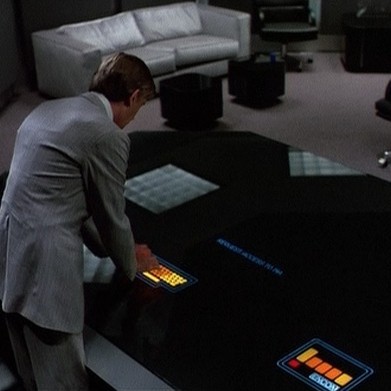 The CEO of the company Encom typing to the console of the MCP from the 1982 movie Tron. He is in an elegant office using a touchscreen interface built into his black glass desk. He wears a grey suit. As he types, he seems to be bowing before the AI he programmed to be ambicious and that started to blackmail him and seized the control.