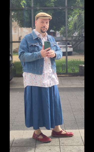 Selbstporträt in spiegelnder Scheibe. Ich trage einen langen weiten Jeansrock, ein weißes Hemd mit bunten Punkten, eine kurze Jeansjacke, eine braune Schirmmütze und rote Schuhe mit T-Spange. Das Hemd hängt lang über den Rock.