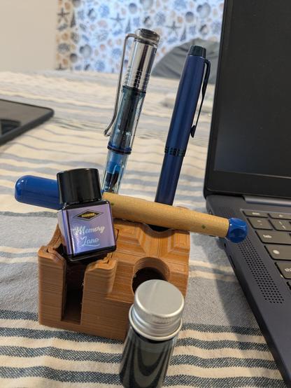 Pen and ink stand with a bottle of Memory Lane ink, an ABC, a LAMY Demonstrator, and an all blue Parker. In front is a bottle of dilute Cosmic Glow.
