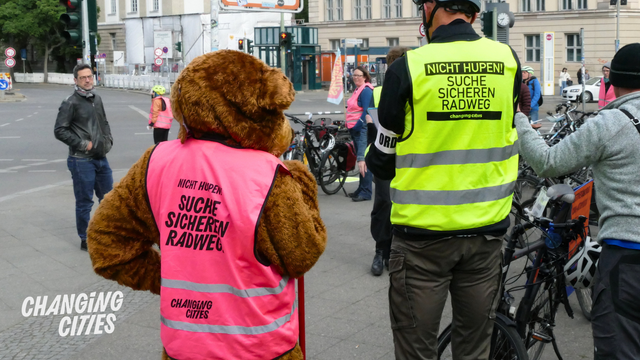 Bär und Mensch nebeneinander: Beide tragen eine Weste: Suche sicheren Radweg.