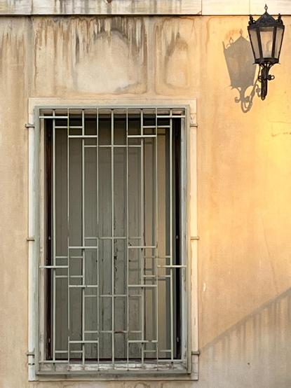 Portrait orientation colour photo of a rectangular window, of almost identical proportions to the image itself, in the lower left of the frame. The window has a thin dingy white marble surround, dingy white closed shutters, and is fronted by a slightly brighter white metal grille which has a geometric vaguely Art Deco-esque pattern of lines and rectangles. The wall in which the window is set is painted a pale yellowish colour, but is waterworn and stained with steaks and blotches of beige and grey. Attached to the wall in the top right hand corner is a small ornate black metal streetlight, its curves and crenellations in contrast to the straight lines of the window. The lamp itself is unlit, but a flood of warm yellow light shines from the right from a source out of the frame, casting a strong shadow from the streetlight, and again emphasising the contrast between the softer curvier lines and the colder rigidness of the window.