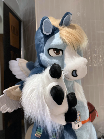 This is a candid waist-up photograph of a blue pegasus fursuiter standing near the entryway to the ConFuzzled Hilton. He's pointing at the camera JoJo-style with a considerable amount of dramatic foreshortening.