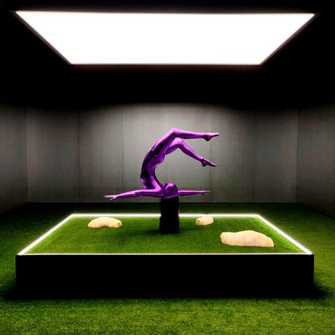 An art installation where a somervaulting woman is in suspended motion in a way that appears she is upside down and her hair is touching the astroturfed square platform. Surrounding her on the platform are three large pale pinkish maggots.