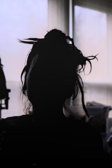 silhouette of a woman's face and upper torso. Her face and head are backlit by a bright window. Her hair is tendrils caught in a messy bun, with sinuous pieces sticking and falling out. One side of her face is slightly illuminated, temple to jawline the rest is in shadow.