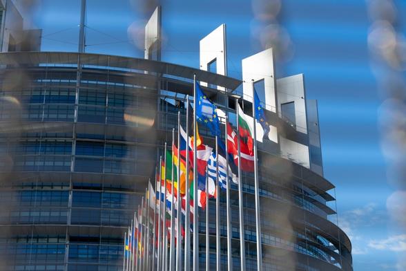 Mehrere Flaggen europäischer Länder und der Europäischen Union wehen vor einem modernen, runden Glasgebäude mit mehreren Stockwerken und markanten weißen Fassadenelementen. Das Gebäude steht vor blauem Himmel, im Vordergrund sind unscharfe, helle Reflexionen sichtbar.