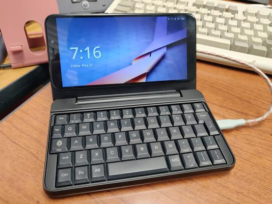 Picture of my PinePhone attached to the PinePhone Keyboard and showing the login screen for Plasma Mobile on postmarketOS. The time is shown on the left side of the screen, and a USB-C cable is attached to the USB-C charging port of the keyboard.