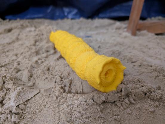 Ein gelber, 3D-gedruckter Sandwurm aus Dune liegt im Sand. Er ist rund im Querschnitt und hat vorne eine Art Maul.